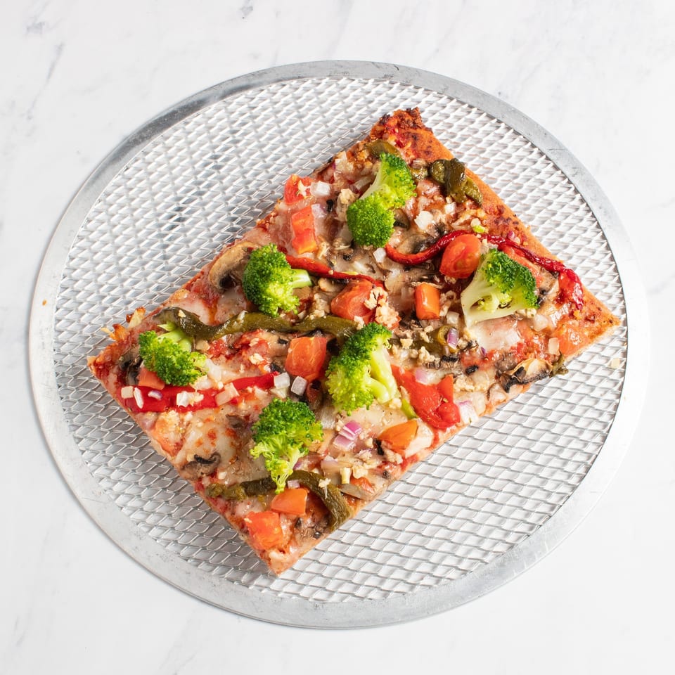 A square slice of pizza with broccoli, tomatoes, onions, and mushrooms on a metal pizza screen.