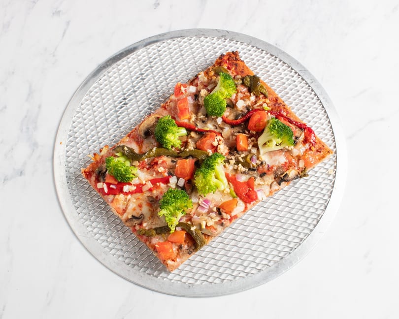 A square slice of pizza with broccoli, tomatoes, onions, and mushrooms on a metal pizza screen.