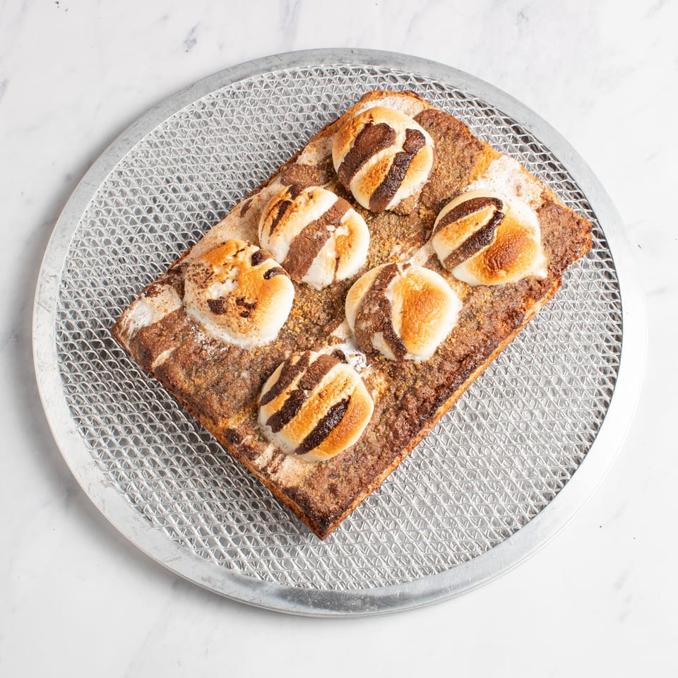 S'mores dessert pizza with toasted marshmallows and chocolate drizzle on a metal pizza screen.