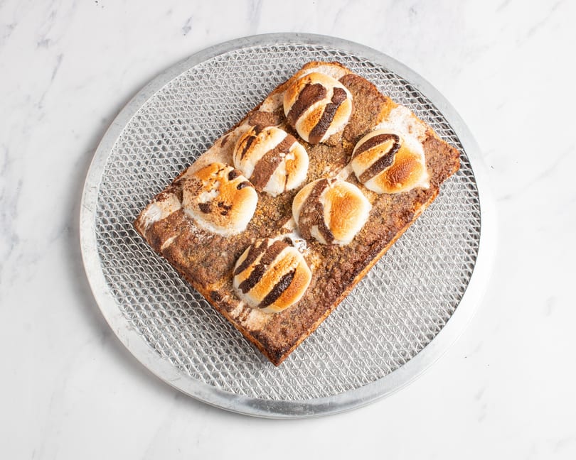 S'mores dessert pizza with toasted marshmallows and chocolate drizzle on a metal pizza screen.