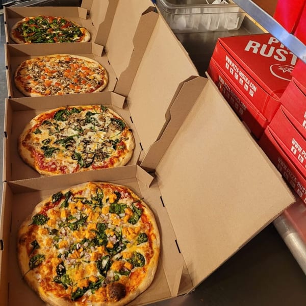 Four Pizza Rustica pizzas in open boxes, with red Pizza Rustica boxes stacked nearby.