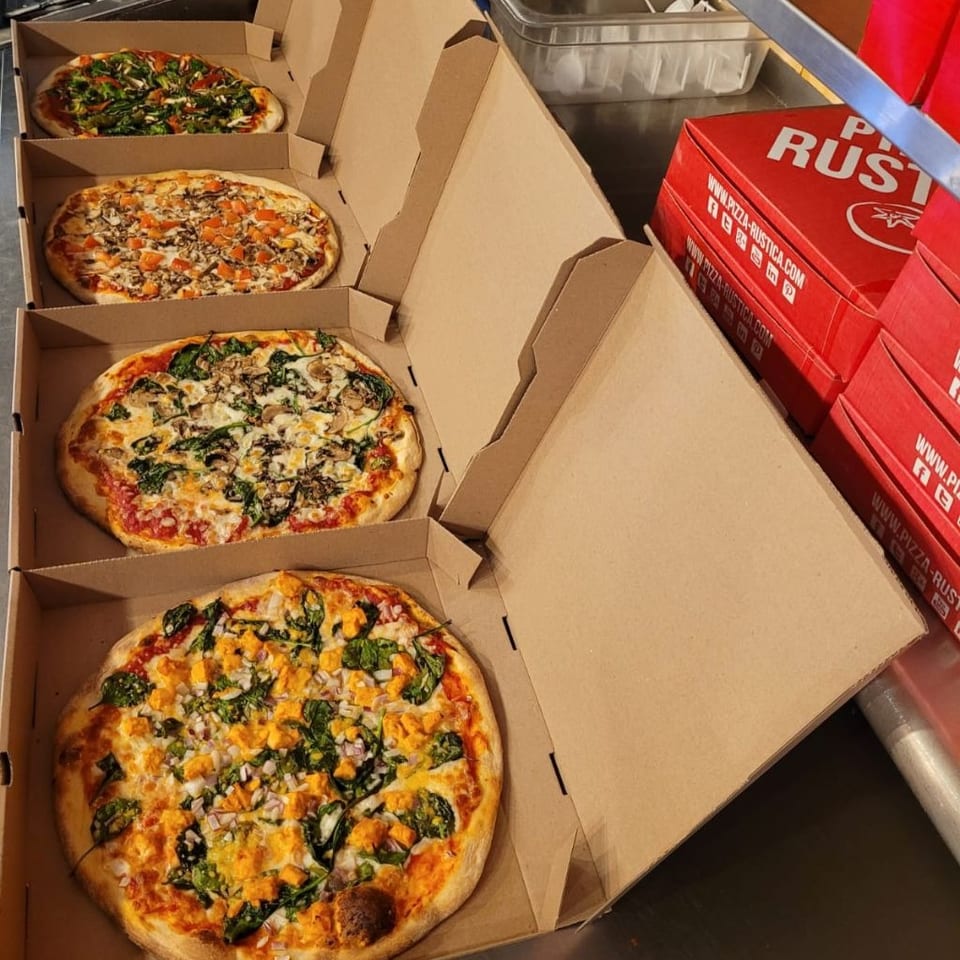 Four Pizza Rustica pizzas in open boxes, with red Pizza Rustica boxes stacked nearby.