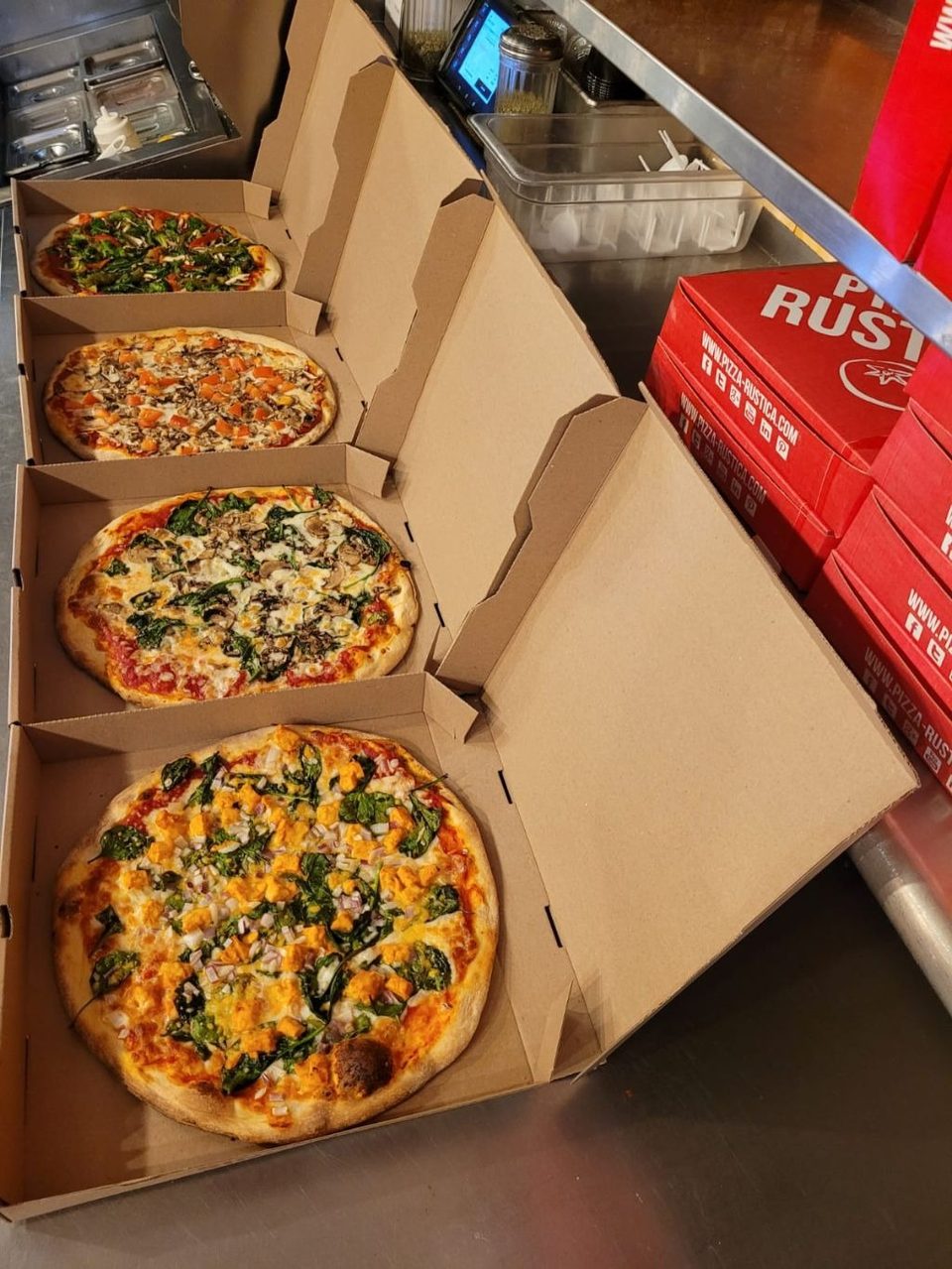 Four Pizza Rustica pizzas in open boxes, with red Pizza Rustica boxes stacked nearby.