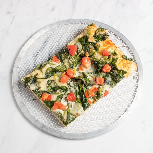 A square slice of pizza with spinach, diced tomatoes, and melted cheese on a metal pizza screen.