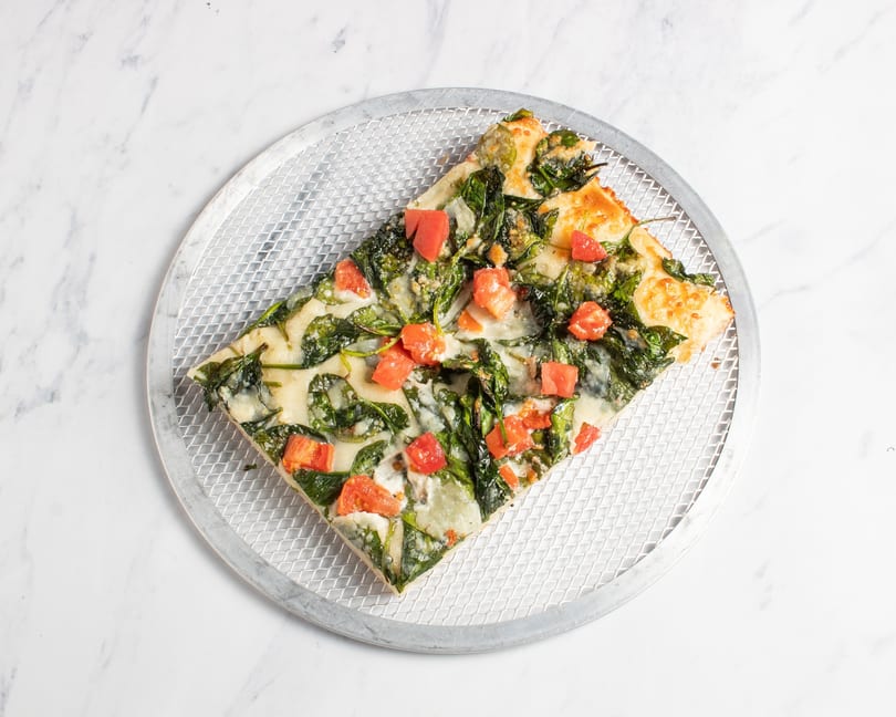 A square slice of pizza with spinach, diced tomatoes, and melted cheese on a metal pizza screen.