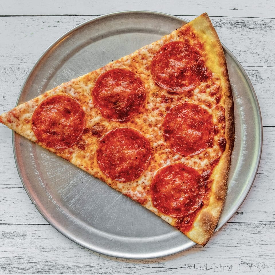 A single slice of pepperoni pizza on a metal tray, viewed from above on a white wooden table.