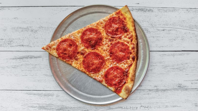 A single slice of pepperoni pizza on a metal tray, viewed from above on a white wooden table.