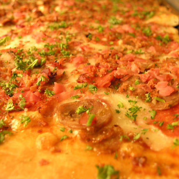Close-up of a rectangular pizza topped with cheese, ham, bacon bits, and fresh parsley.