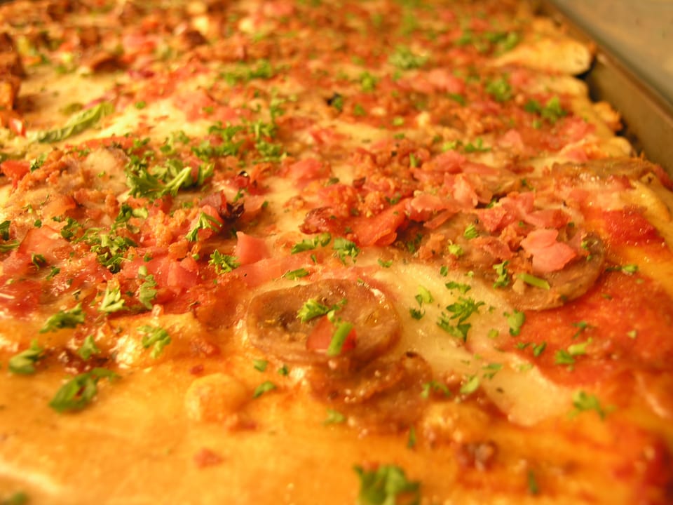 Close-up of a rectangular pizza topped with cheese, ham, bacon bits, and fresh parsley.